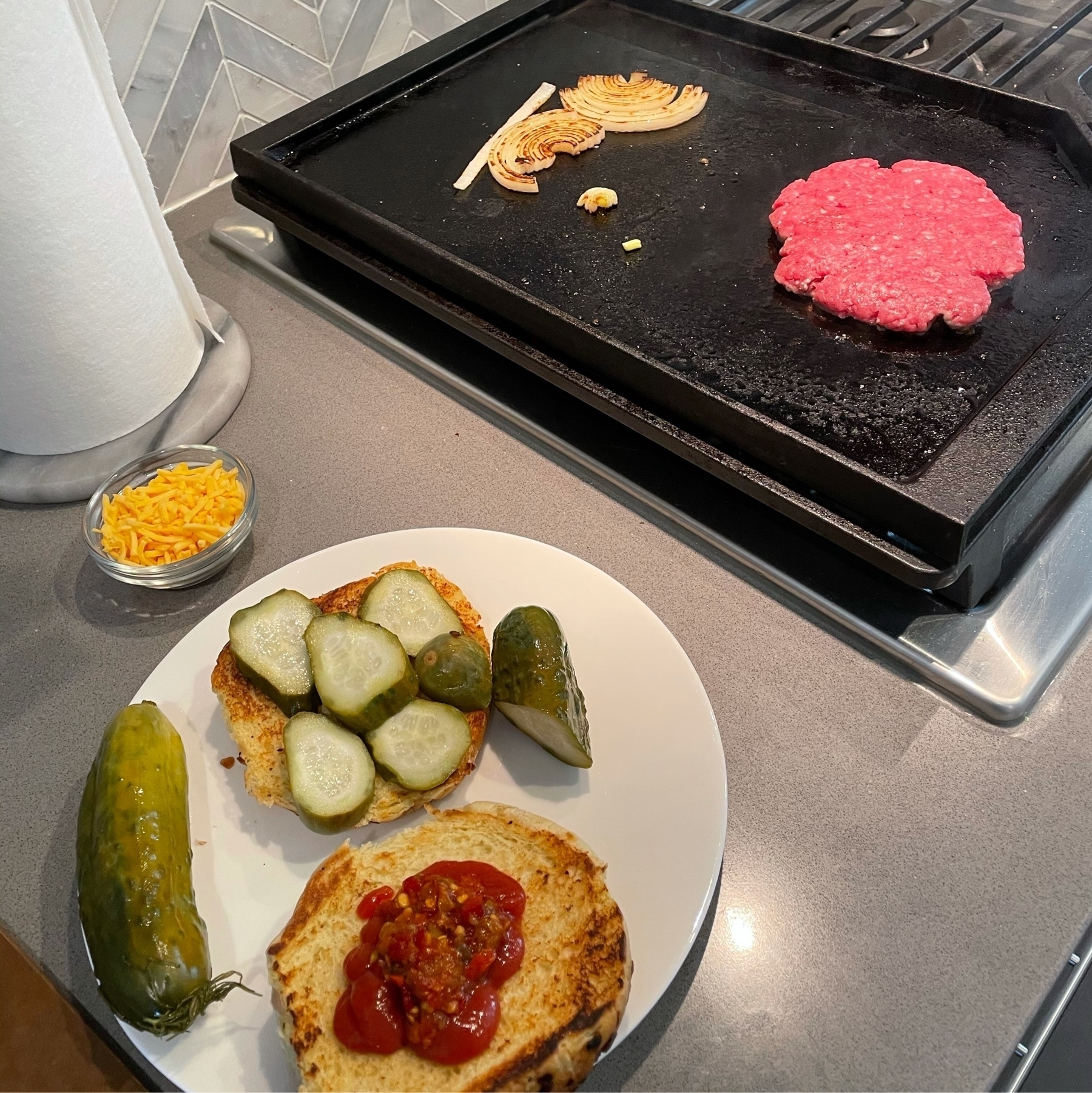 Burger on a flat top, with onions grilling, and a bun already assembled with pickles and ketchup.