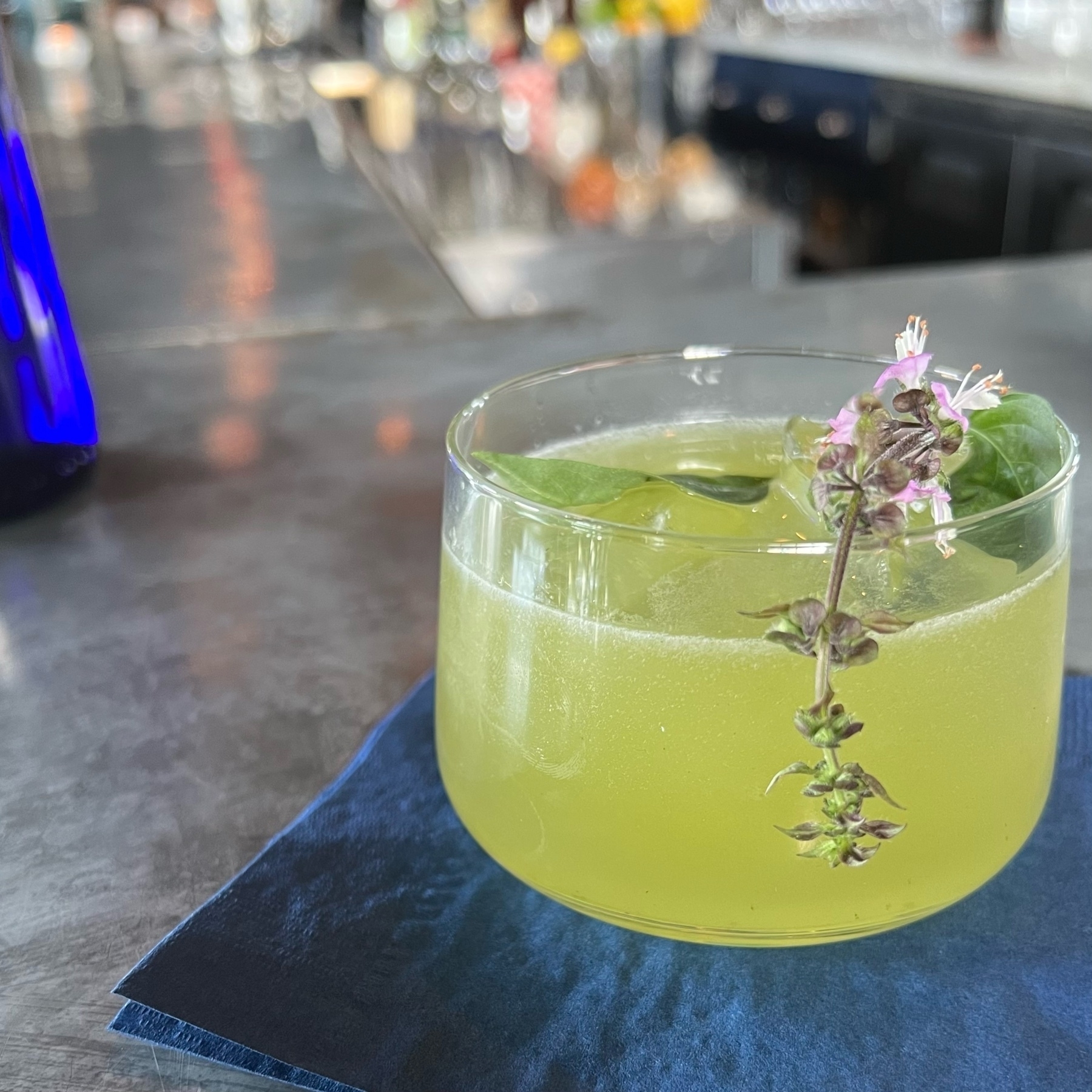 Green cocktail on a bar with a purple flour and basil leaf