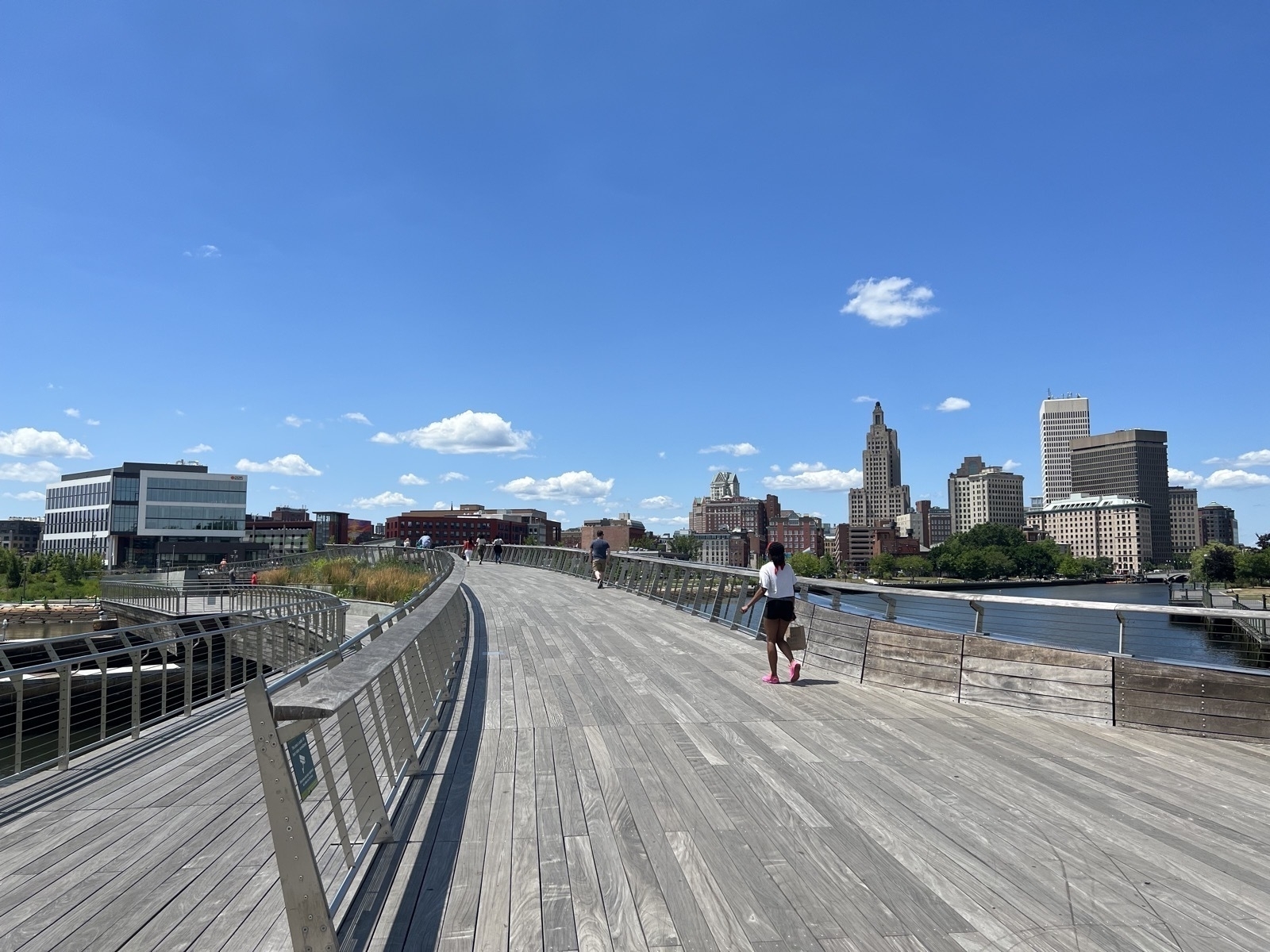 A light gray, wooden, pedestrian bridge with steel railings over the water in Providence RI. The city skyline and park are in the background