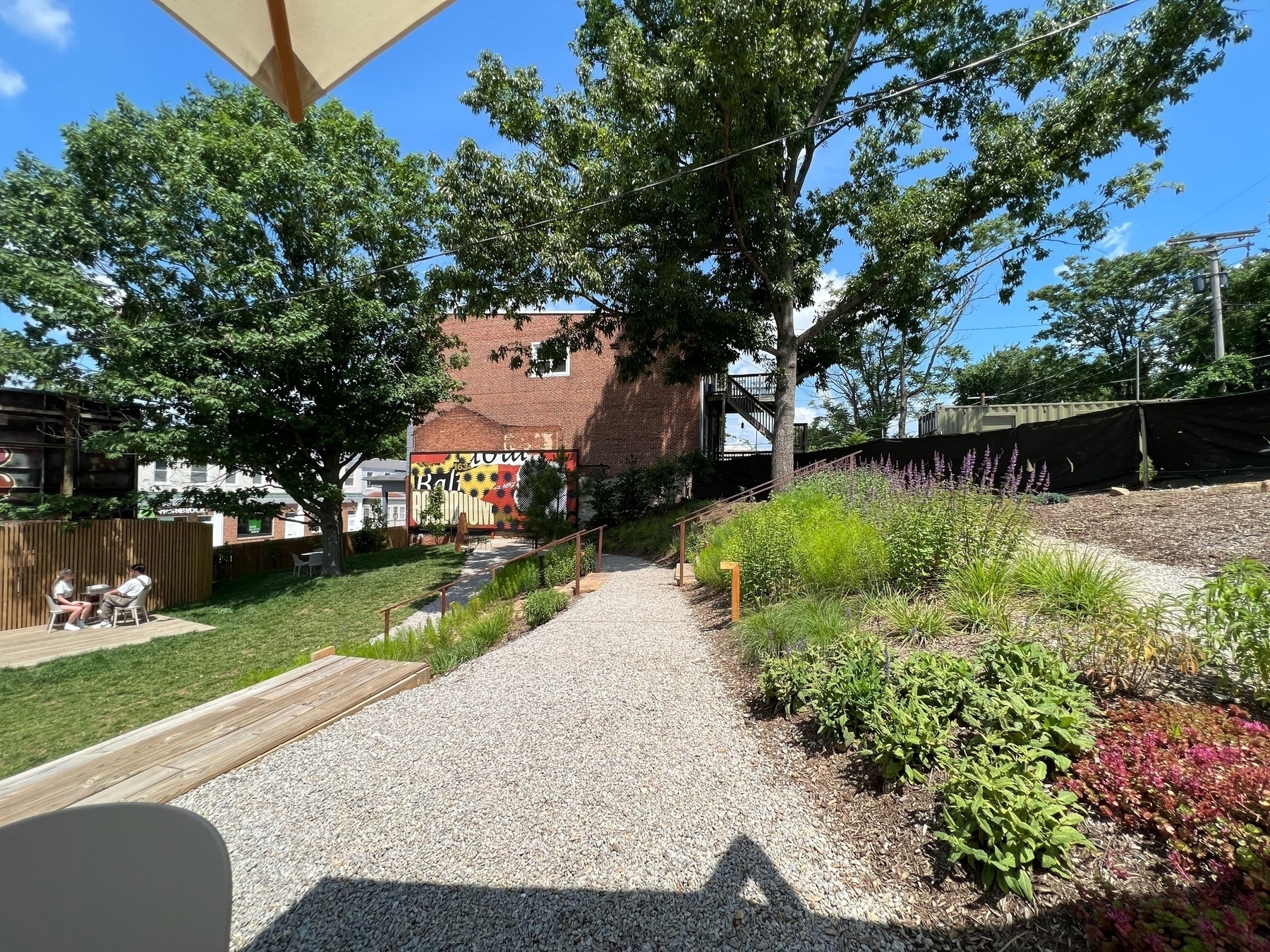 Outdoor view at good neighbor coffee shop, which is a terraced garden with gravel and wood steps in the foreground and a small lawn and tree in the background. There&rsquo;s also a Baltimore billboard on the property against a brick wall.