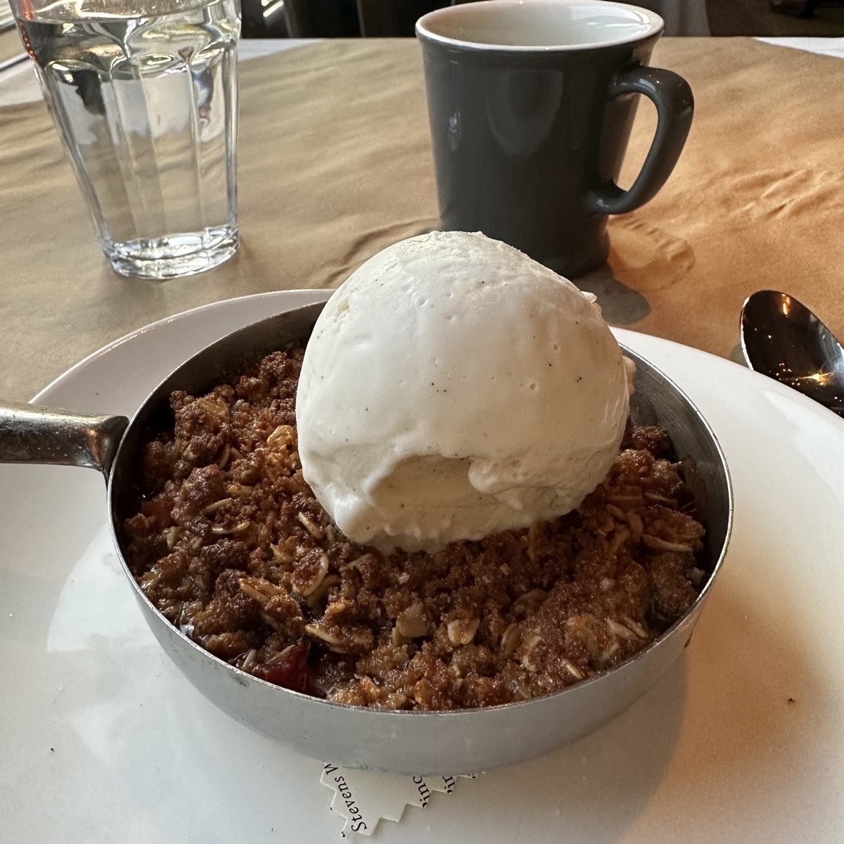 A large ball of ice cream atop a rhubarb crumble in a round aluminum bowl with a handle.