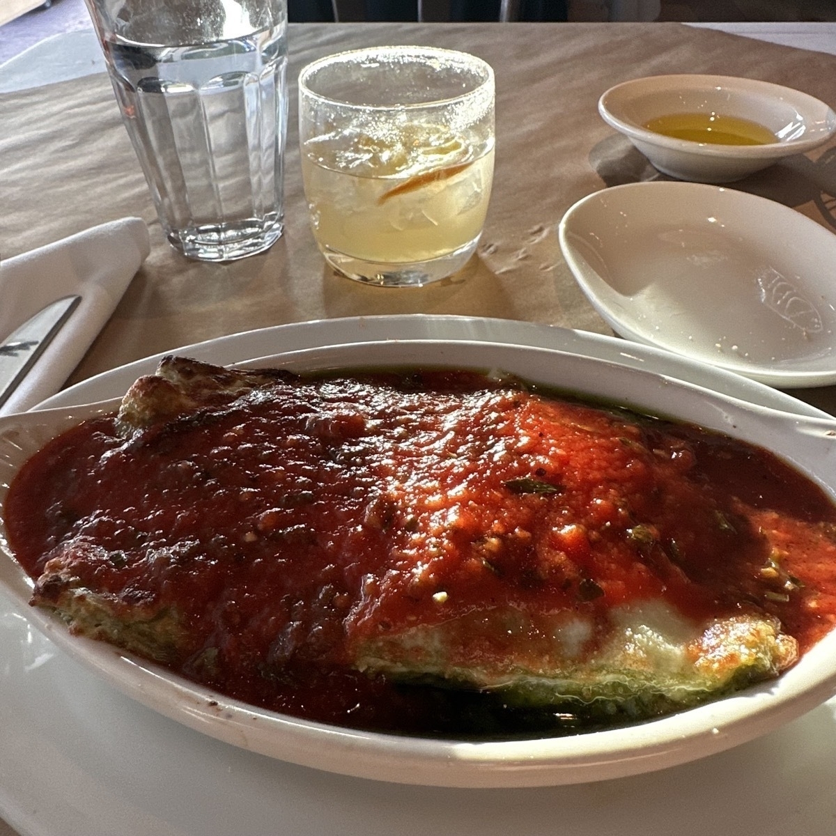 Lasagna in a boat shaped porcelain white plate, with a yellow drink behind.