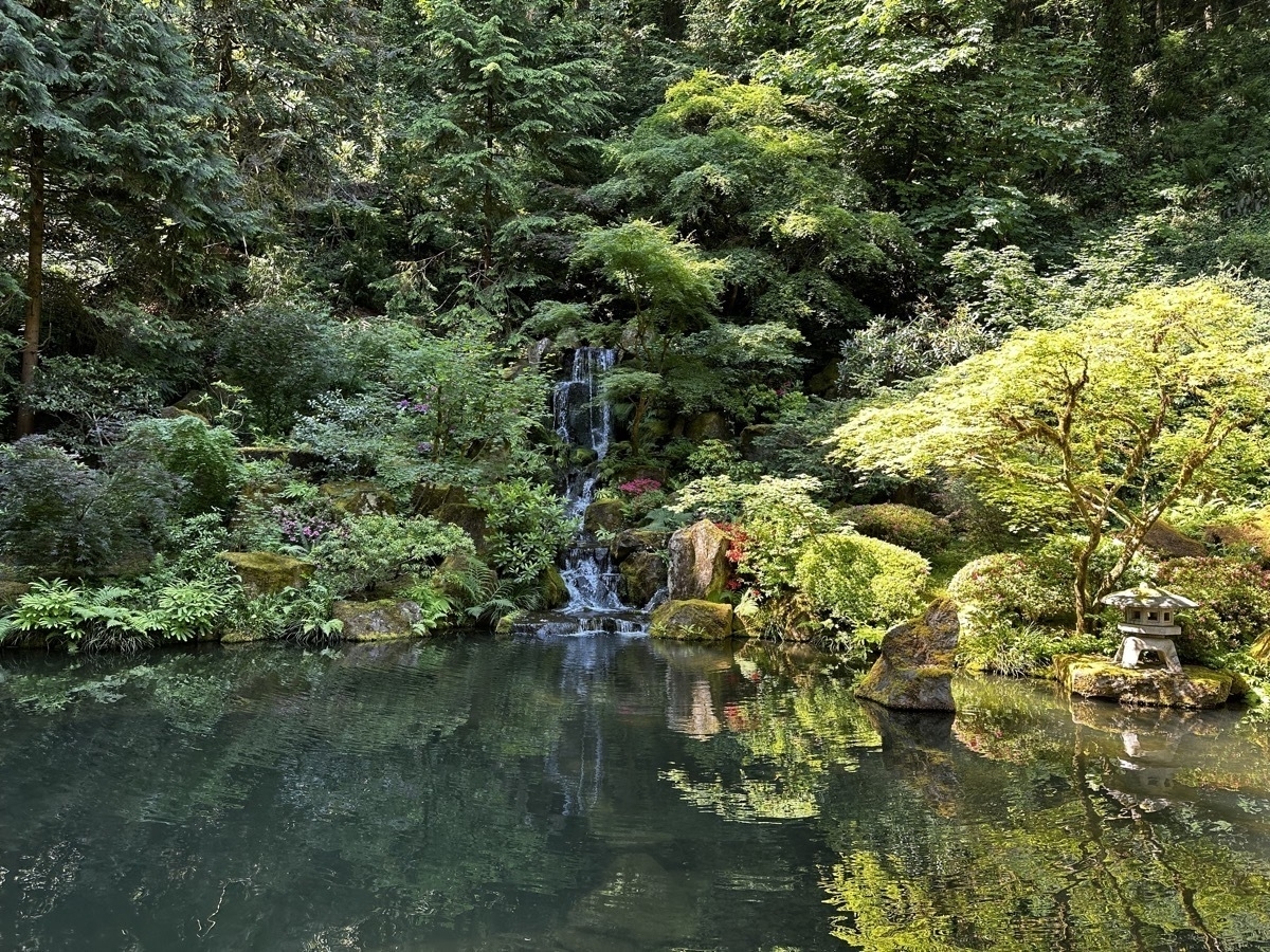 A lake surrounded by rich greenery, with a small waterfall in the background and a small stone hut in the foreground to the left at Japanese Garden in Portland, Oregon.